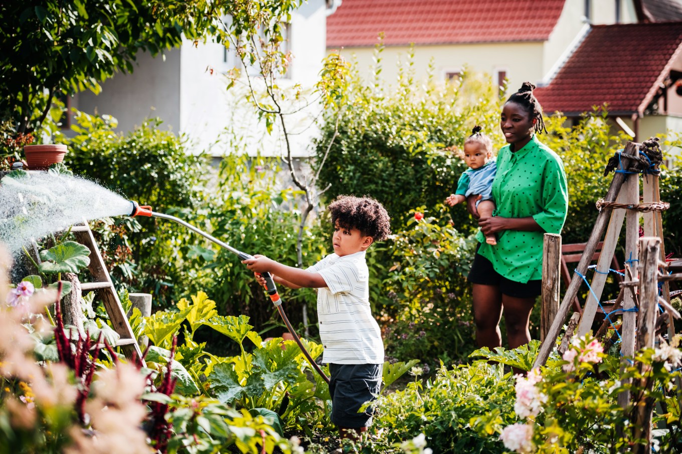 A young boy manually watering flowers in a garden being guided by his mom.