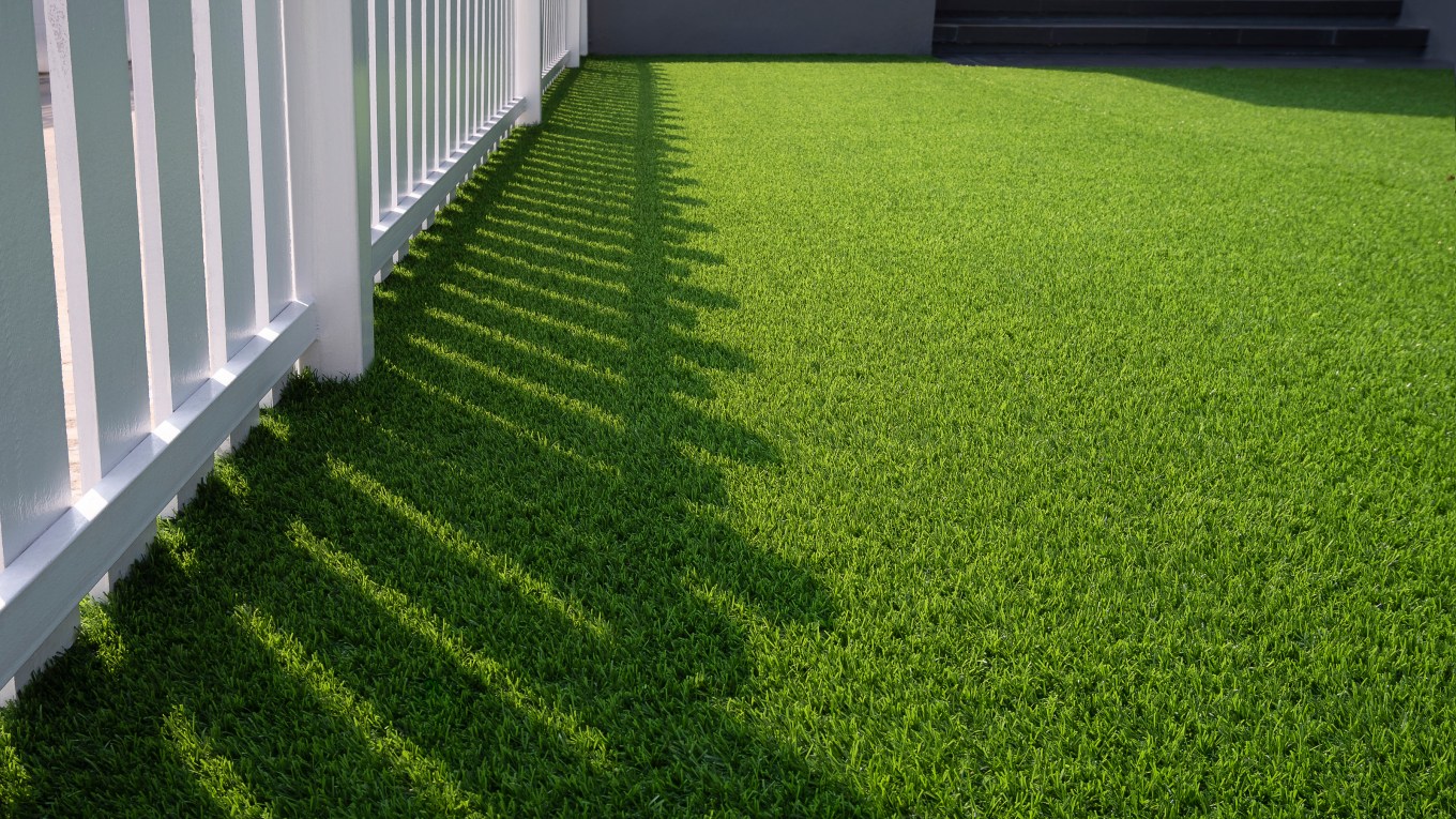 A white wooden fence casts a shadow on green artificial grass that's UV protected.