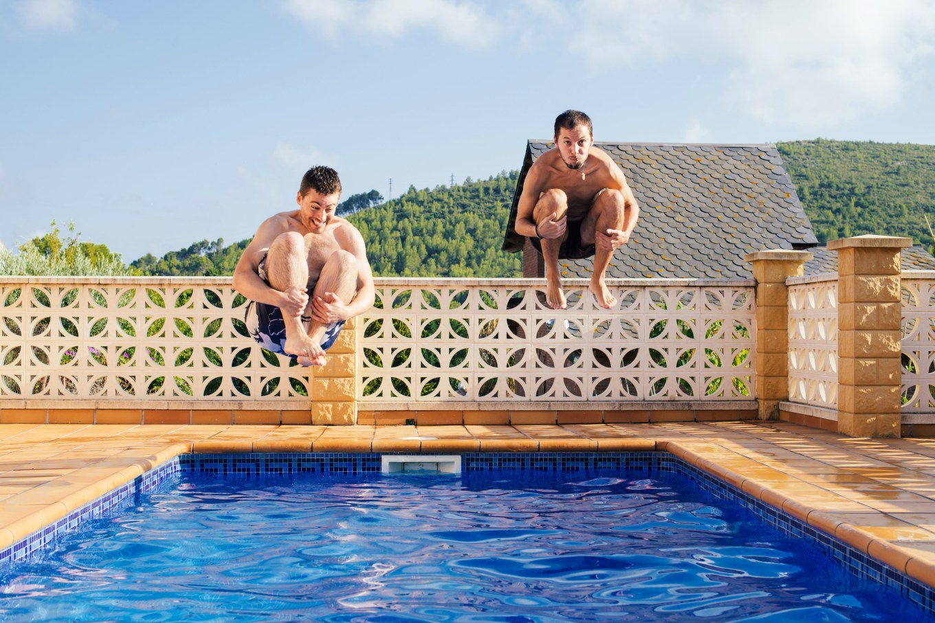 Two people cannonballing into a pool with a concrete tile fence.