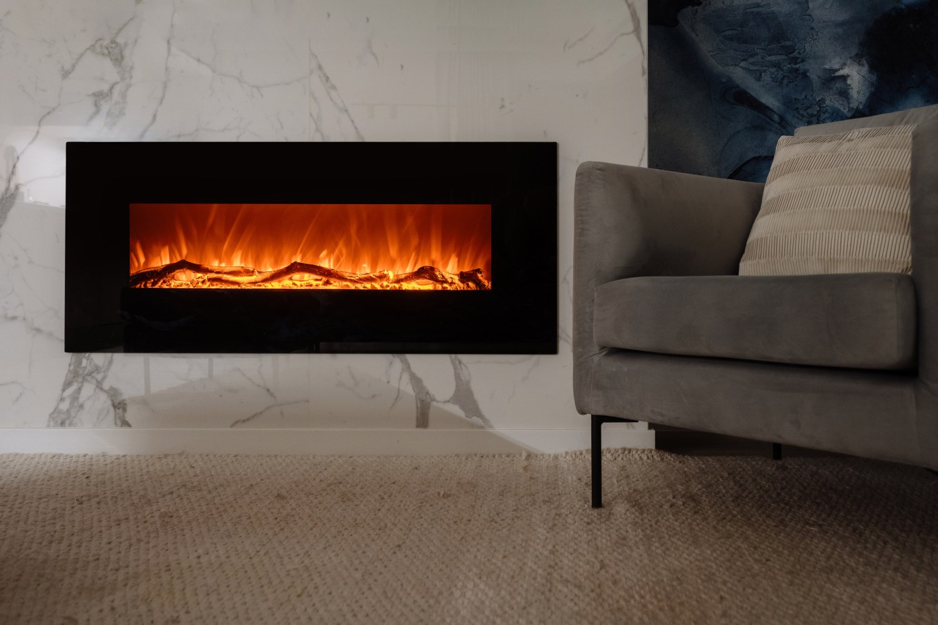 Modern electric fireplace with realistic flames mounted on white marble wall. Cozy gray chair sits near fireplace in living room. Quiet evening in hotel or weekend at home