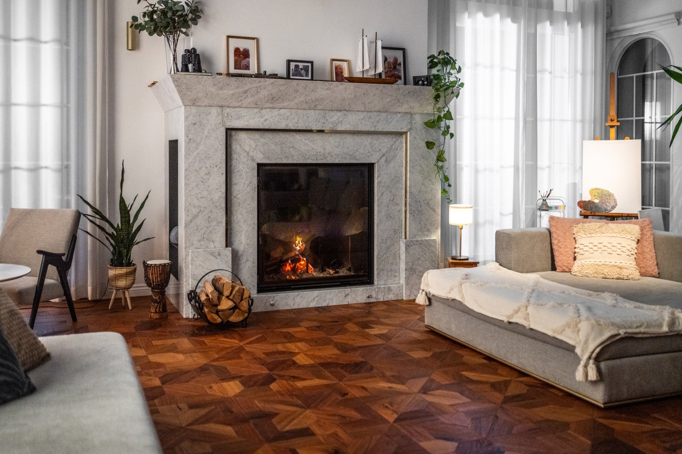 Spacious living room showcases a roaring fire within a prominent fireplace, flanked by comfortable furniture and tasteful decorations on a herringbone wood floor.